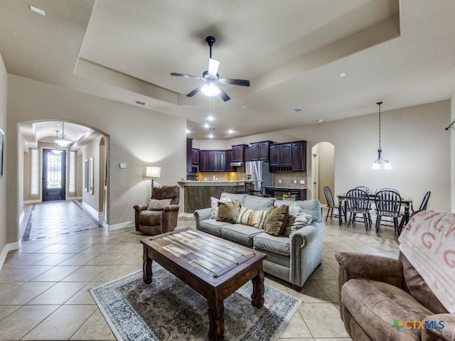 tiled living room with ceiling fan with notable chandelier and a tray ceiling