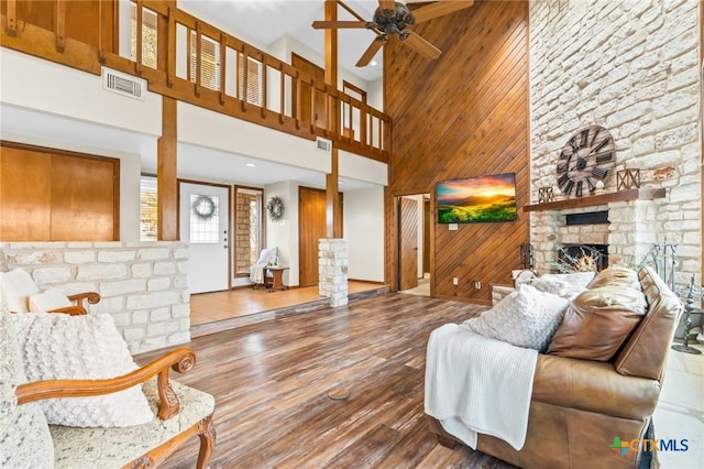 living room with ceiling fan, a high ceiling, a stone fireplace, wood walls, and wood-type flooring