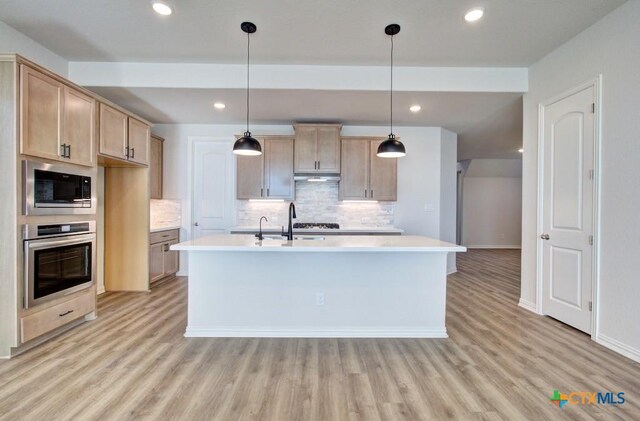 kitchen featuring built in microwave, light brown cabinetry, stainless steel oven, hanging light fixtures, and a center island with sink