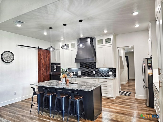 kitchen featuring custom exhaust hood, a barn door, a center island with sink, hardwood / wood-style floors, and stainless steel refrigerator