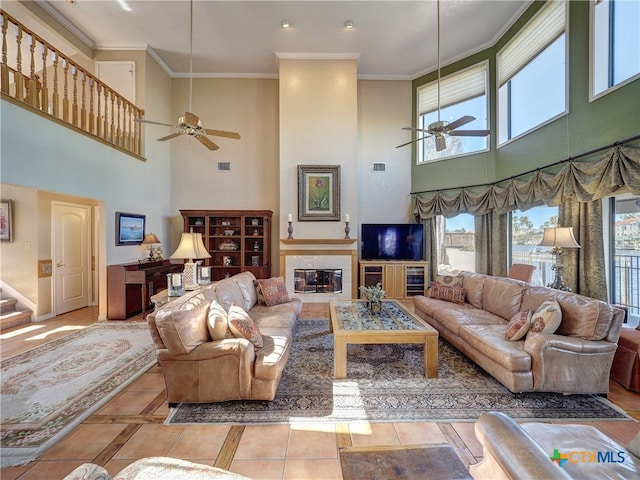 tiled living room with ceiling fan, a towering ceiling, crown molding, and a fireplace