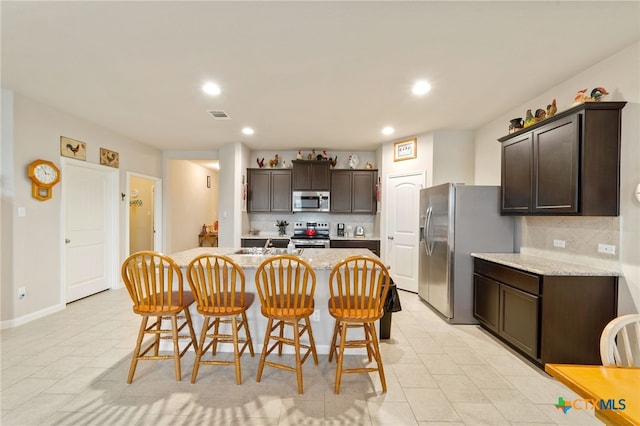 kitchen with light stone countertops, stainless steel appliances, a kitchen breakfast bar, backsplash, and an island with sink