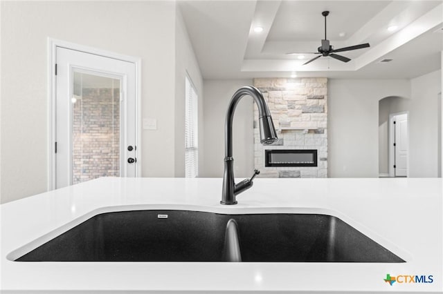 kitchen with ceiling fan, a tray ceiling, sink, and a stone fireplace