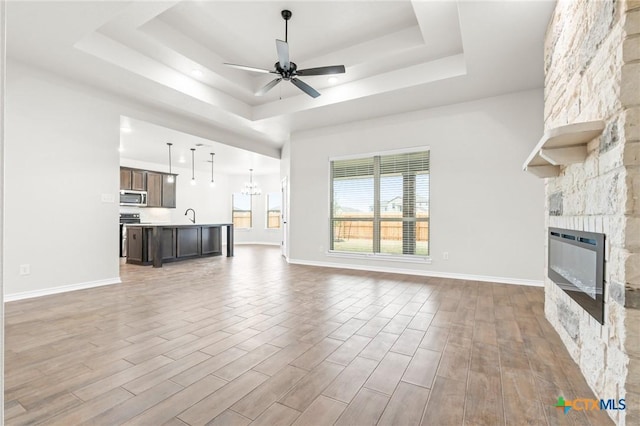 unfurnished living room featuring sink, a tray ceiling, ceiling fan with notable chandelier, and a stone fireplace