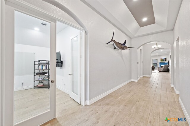 hall with a raised ceiling, light wood-style flooring, baseboards, visible vents, and arched walkways