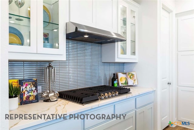 kitchen with under cabinet range hood, stainless steel gas cooktop, white cabinetry, decorative backsplash, and glass insert cabinets