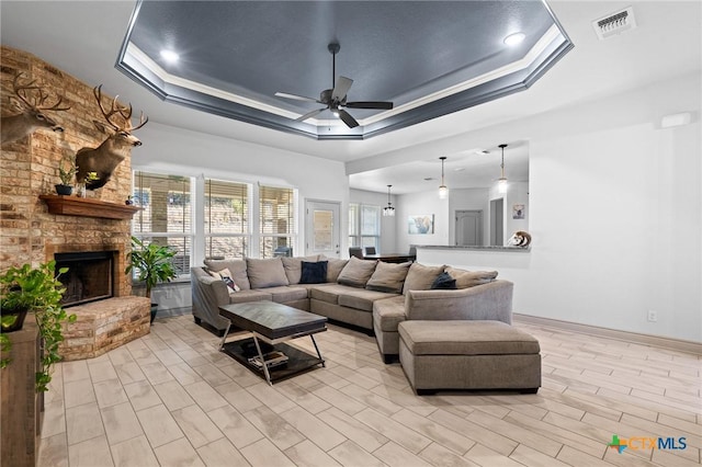 living room with ceiling fan with notable chandelier, a tray ceiling, a brick fireplace, and crown molding