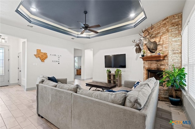 tiled living room with a brick fireplace, ceiling fan, crown molding, and a tray ceiling