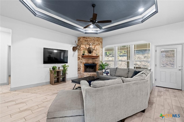 living room with a tray ceiling, a stone fireplace, ceiling fan, and ornamental molding