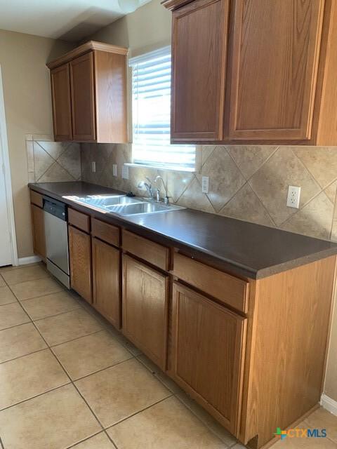 kitchen featuring a sink, backsplash, stainless steel dishwasher, dark countertops, and light tile patterned floors