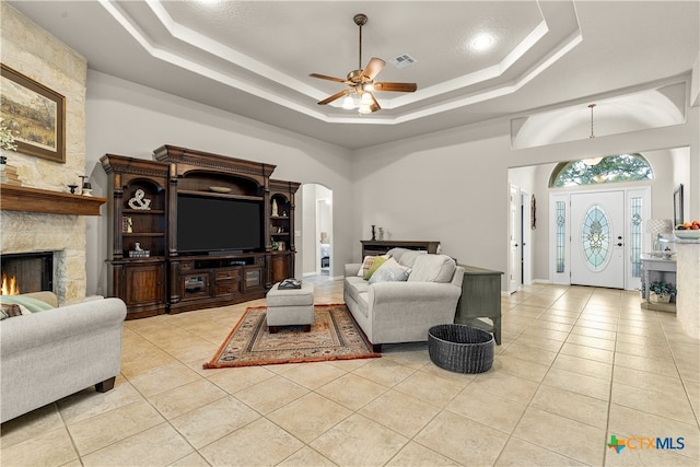 living room featuring a stone fireplace, light tile patterned flooring, ceiling fan, and a tray ceiling