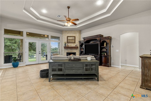 tiled living room featuring ceiling fan, a stone fireplace, french doors, and a tray ceiling