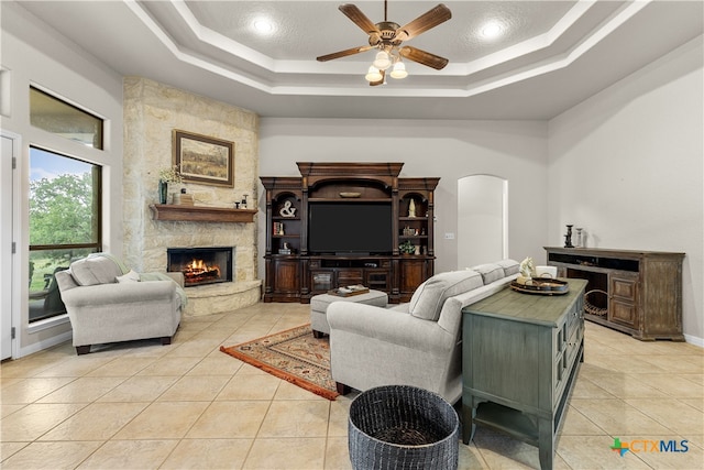 living room featuring a stone fireplace, ceiling fan, a tray ceiling, and light tile patterned floors