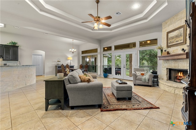 tiled living room featuring french doors, a stone fireplace, a raised ceiling, sink, and ceiling fan with notable chandelier