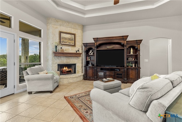 tiled living room with ceiling fan, a stone fireplace, and a tray ceiling