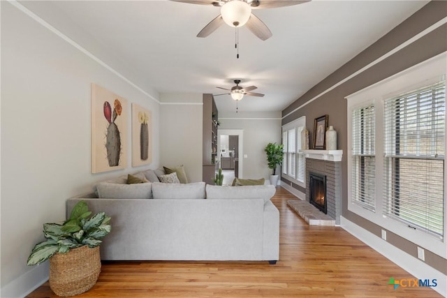 living room featuring light wood-style floors, baseboards, a ceiling fan, and a brick fireplace
