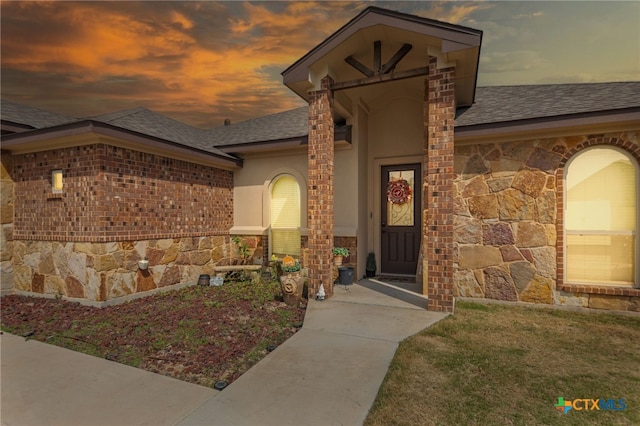exterior entry at dusk with stone siding, a shingled roof, brick siding, and stucco siding