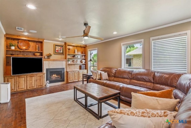 living room featuring wood finished floors, visible vents, crown molding, a tiled fireplace, and ceiling fan