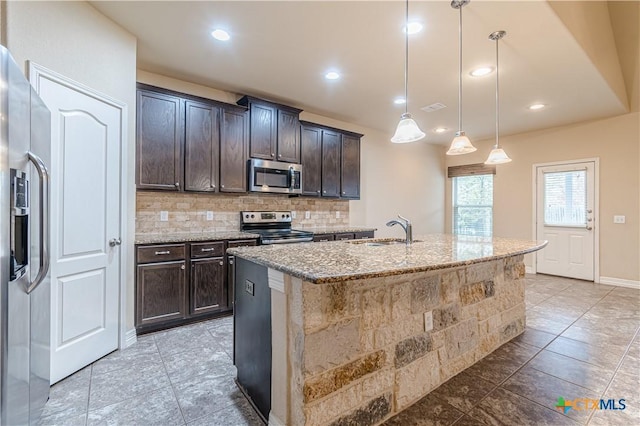 kitchen featuring sink, light stone counters, hanging light fixtures, appliances with stainless steel finishes, and a kitchen island with sink