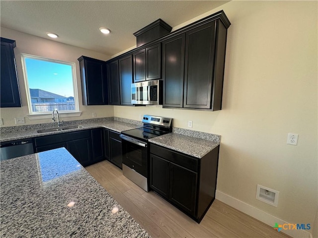 kitchen with light stone counters, light wood finished floors, stainless steel appliances, a sink, and dark cabinets