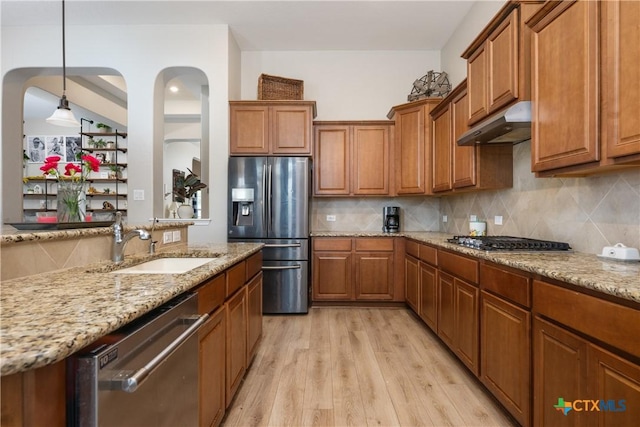 kitchen featuring a sink, under cabinet range hood, decorative backsplash, brown cabinetry, and appliances with stainless steel finishes