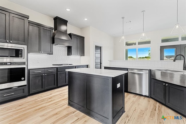 kitchen with appliances with stainless steel finishes, decorative light fixtures, light hardwood / wood-style flooring, a kitchen island, and premium range hood