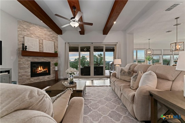 living room with beamed ceiling, plenty of natural light, a stone fireplace, and ceiling fan with notable chandelier