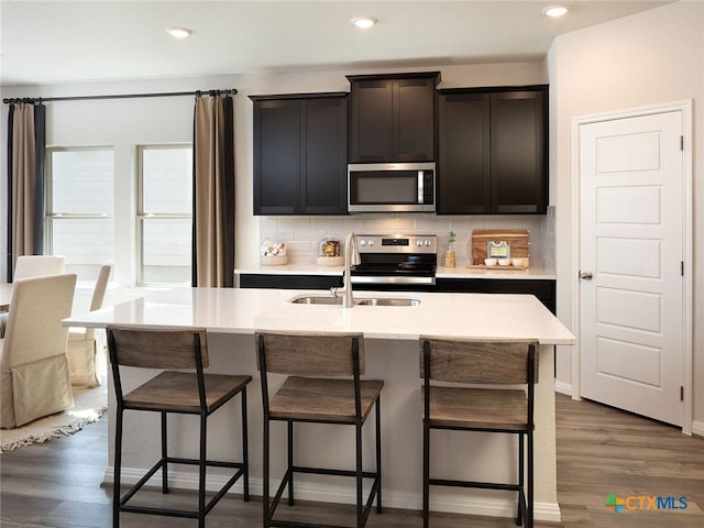 kitchen with dark hardwood / wood-style flooring, sink, tasteful backsplash, an island with sink, and appliances with stainless steel finishes