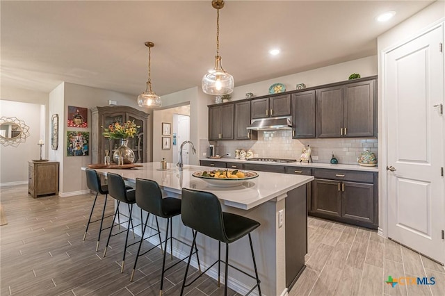 kitchen with light wood-type flooring, under cabinet range hood, stainless steel gas cooktop, a kitchen breakfast bar, and dark brown cabinetry