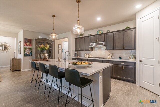 kitchen featuring decorative light fixtures, a kitchen bar, stainless steel gas cooktop, a kitchen island with sink, and dark brown cabinets