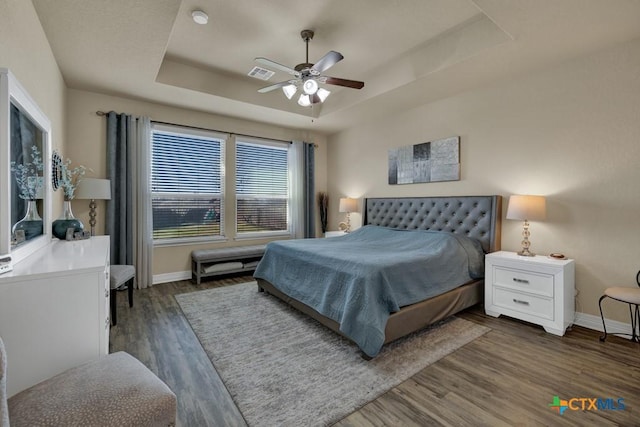 bedroom featuring wood finished floors, visible vents, baseboards, a tray ceiling, and ceiling fan