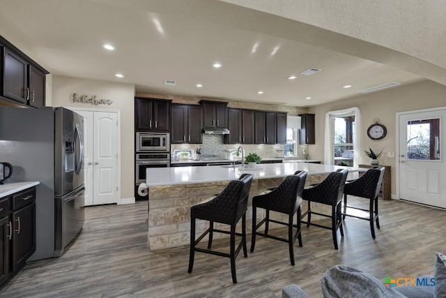 kitchen featuring wood finished floors, decorative backsplash, under cabinet range hood, appliances with stainless steel finishes, and a kitchen breakfast bar