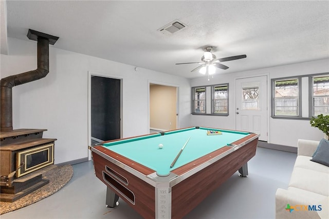 rec room featuring finished concrete flooring, visible vents, a wood stove, ceiling fan, and a textured ceiling