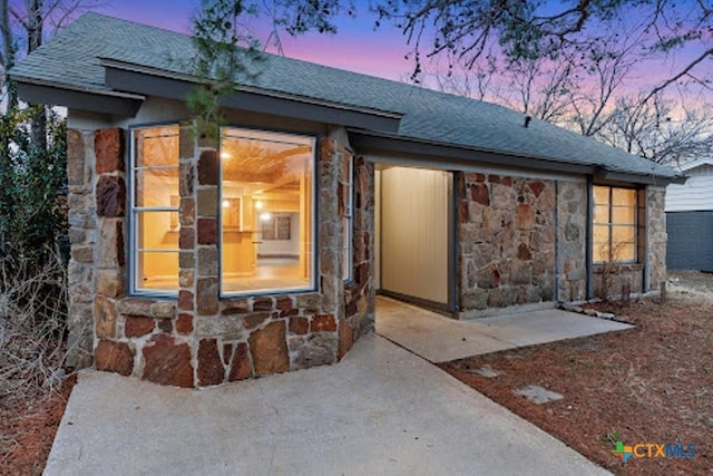 exterior entry at dusk featuring stone siding and roof with shingles