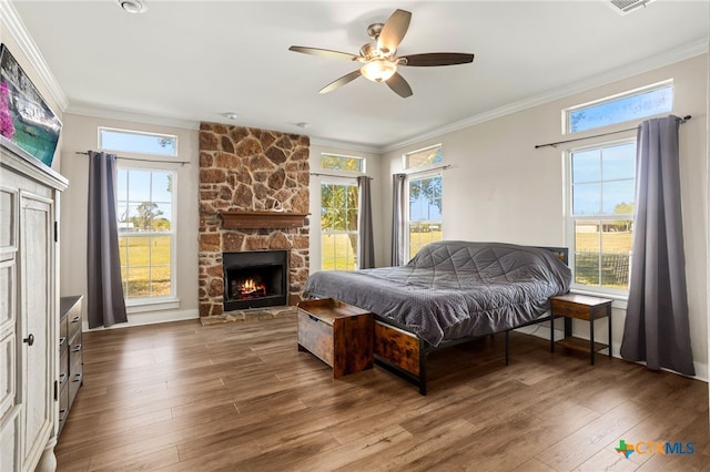 bedroom with a stone fireplace, dark wood-type flooring, multiple windows, and ceiling fan