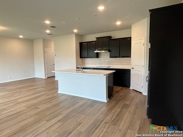 kitchen with tasteful backsplash, black stovetop, sink, light hardwood / wood-style floors, and a kitchen island with sink