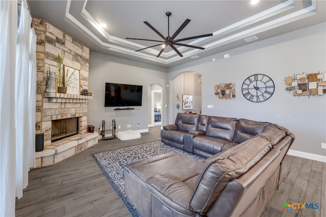 living room with a stone fireplace, a tray ceiling, hardwood / wood-style floors, and ceiling fan