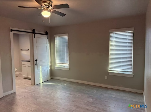 unfurnished bedroom with ceiling fan, a barn door, light wood-type flooring, and ensuite bath