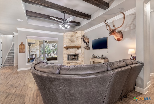 living room featuring beamed ceiling, ornamental molding, ceiling fan, a stone fireplace, and light hardwood / wood-style flooring