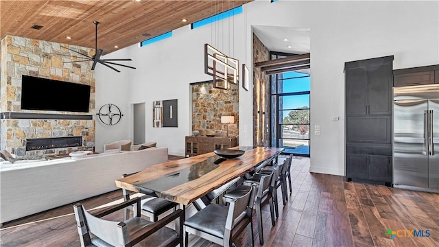 dining room with wood ceiling, a towering ceiling, a stone fireplace, and dark wood-type flooring