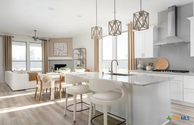 kitchen with a sink, ceiling fan, wall chimney exhaust hood, a center island with sink, and stainless steel gas cooktop