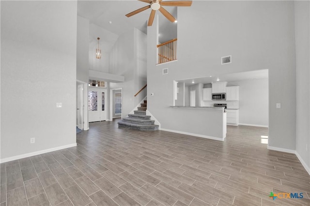 unfurnished living room with a ceiling fan, stairs, a high ceiling, light wood-type flooring, and baseboards