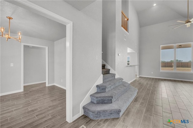 stairs with ceiling fan with notable chandelier, baseboards, wood finish floors, and high vaulted ceiling