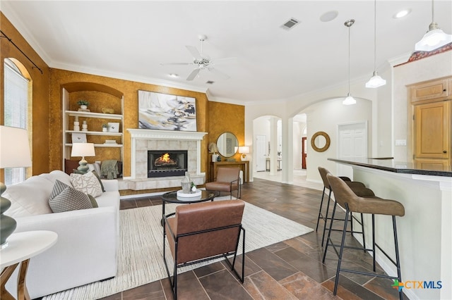living room featuring ceiling fan, arched walkways, built in features, crown molding, and a tiled fireplace
