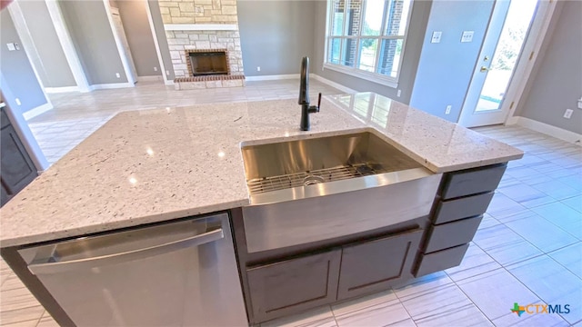 kitchen featuring a stone fireplace, a center island with sink, stainless steel dishwasher, and light stone countertops