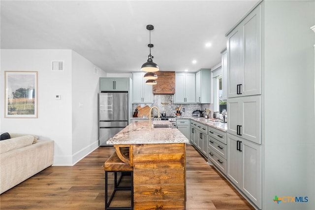 kitchen with pendant lighting, an island with sink, sink, stainless steel fridge, and light stone counters