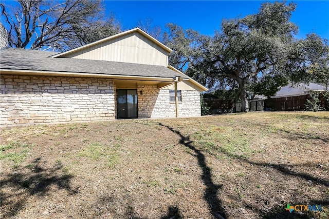 rear view of house featuring stone siding, roof with shingles, fence, and a lawn