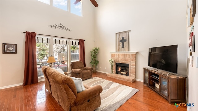 living room featuring ceiling fan, a towering ceiling, wood-type flooring, and a brick fireplace