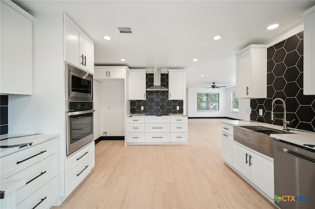 kitchen with stainless steel appliances, ceiling fan, wall chimney range hood, white cabinets, and light hardwood / wood-style floors