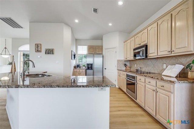 kitchen with stainless steel appliances, a sink, light brown cabinets, a center island with sink, and tasteful backsplash